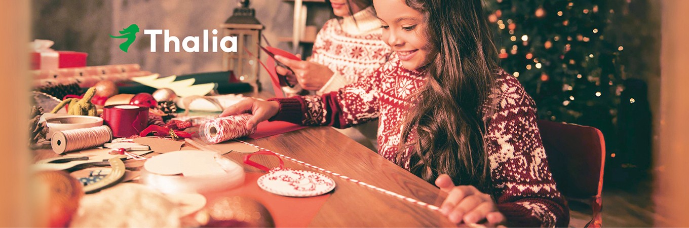 Eine fröhliche Szene in weihnachtlicher Atmosphäre: Ein Mädchen bastelt gemeinsam mit einer Frau an einem festlich geschmückten Tisch. Beide tragen rot-weiße Norwegerpullover. Auf dem Tisch liegen Bastelmaterialien wie Papier, Schnur, Scheren, Schleifen und Dekoration. Im Hintergrund leuchtet ein geschmückter Weihnachtsbaum. Links oben ist das Thalia-Logo mit dem grünen Lesemädchen zu sehen.