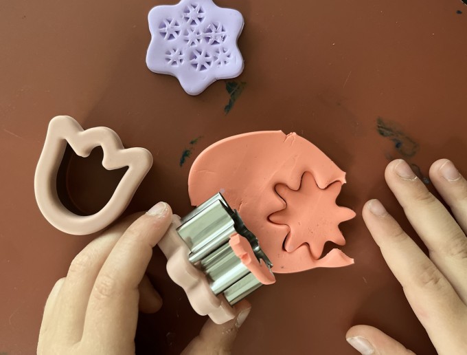 Zwei Kinderhände arbeiten mit rosafarbener Knetmasse auf einer rotbraunen Unterlage. Eine metallene Ausstechform in Form einer gezackten Blume wird gerade in die Knetmasse gedrückt. Daneben liegen weitere Ausstechformen: eine rosa Tulpenform und eine lilafarbene Form mit sternartigen Mustern.