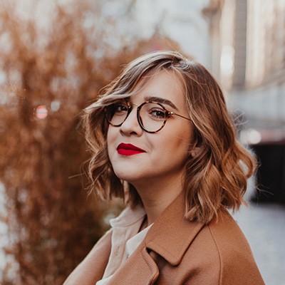 Frau mit hellbraunen schulterlangen Haaren, roten Lippenstift, Brille und brauner Jacke.