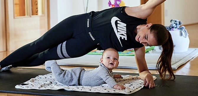 Eine Frau macht auf einer Sportmatte eine Workout Übung. Vor ihr liegt ein kleines Kind.