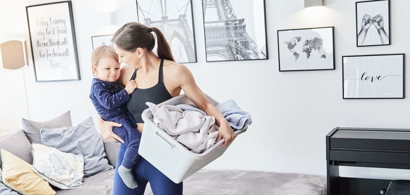 Eine Frau steht im Wohnzimmer. In einer Hand hält sie einen kleinen Jungen, in der anderen einen Wäschekorb.